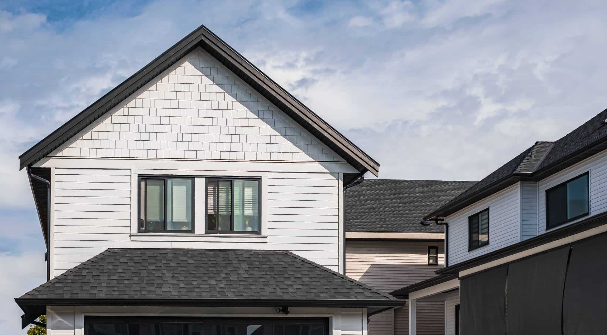 A modern two-story house with light-colored siding, black window frames, and a dark shingle roof, situated next to a similar neighboring home under a partly cloudy sky.