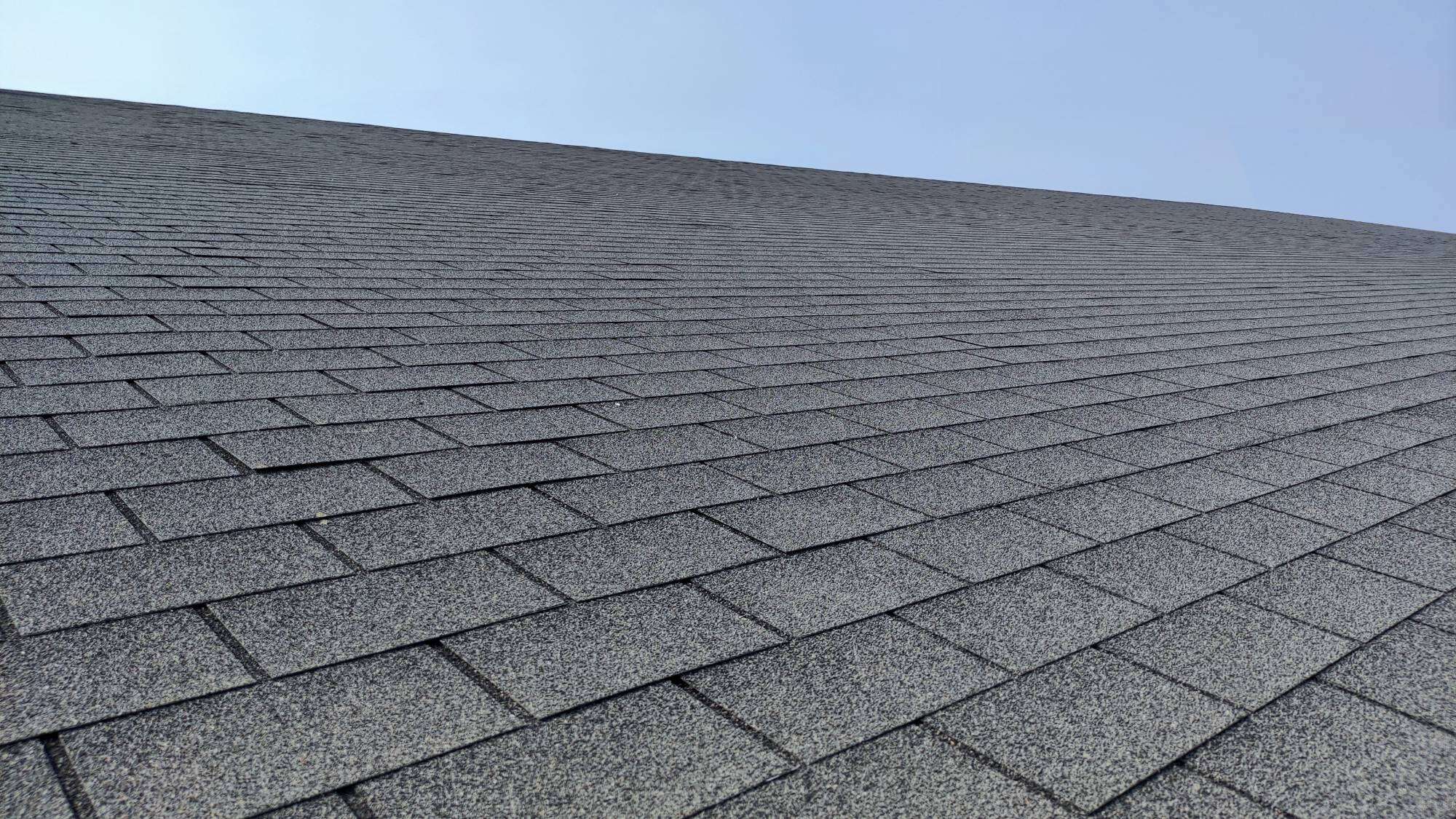 A close-up view looking up at a sloped roof covered with gray asphalt shingles, with a clear blue sky in the background.