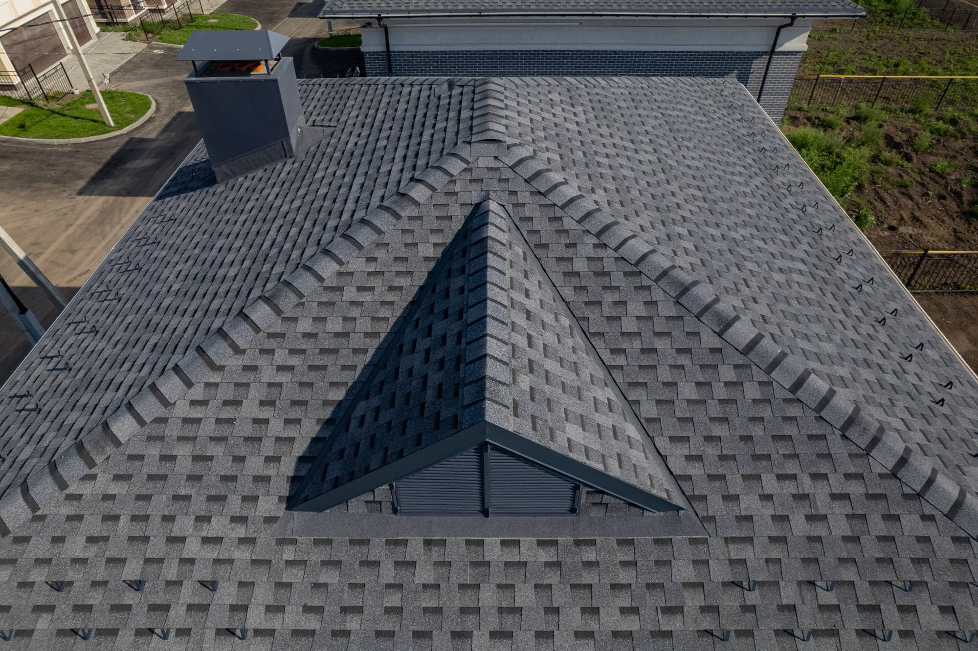Aerial view of a gray shingle roof with a prominent triangular peak and vent, surrounded by a paved area, grass, and adjacent buildings.