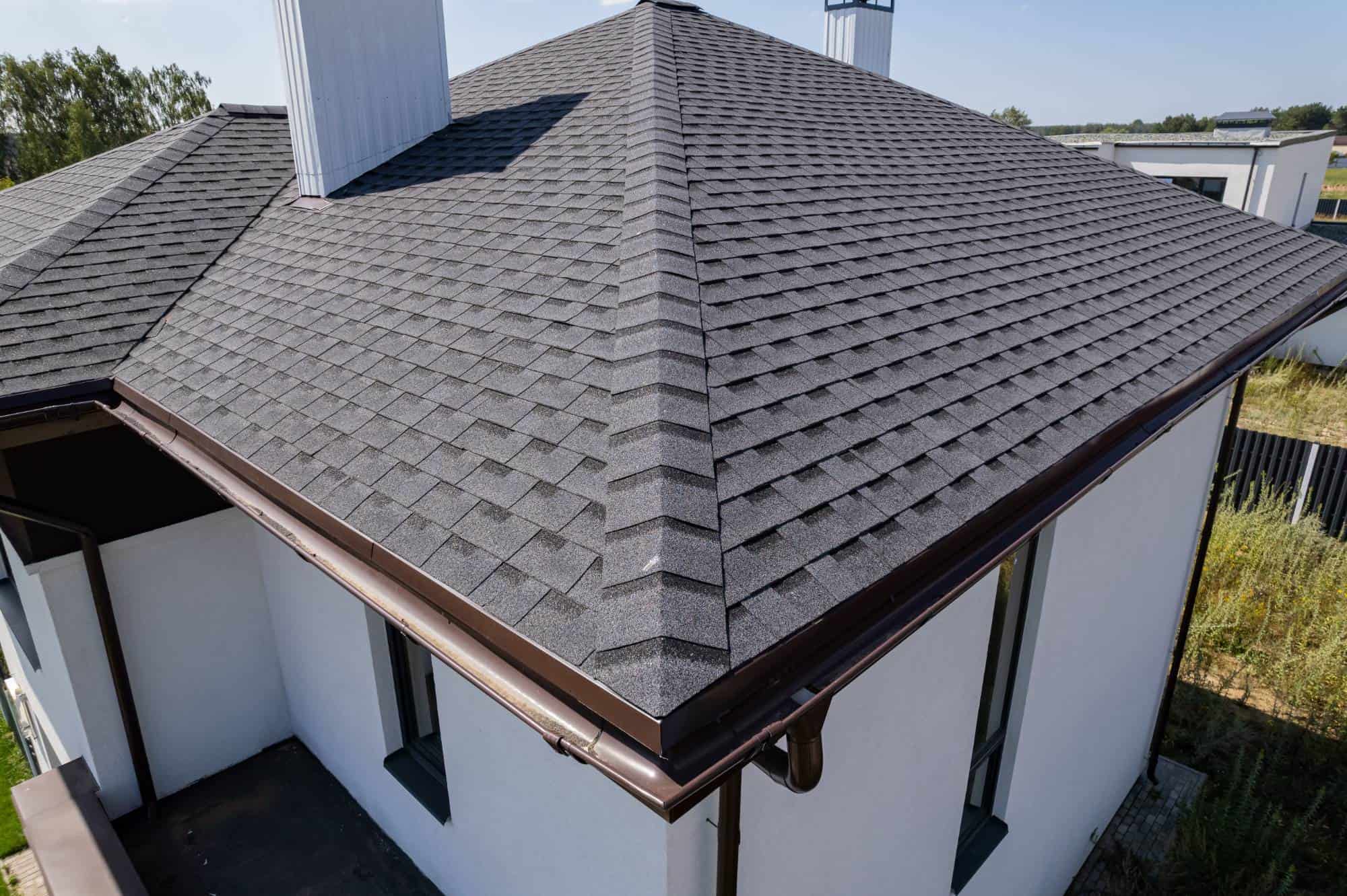 A close-up view of the black shingled roof of a modern white house, showing neat, geometric shingles and clean brown gutters, with greenery and neighboring buildings visible in the background.