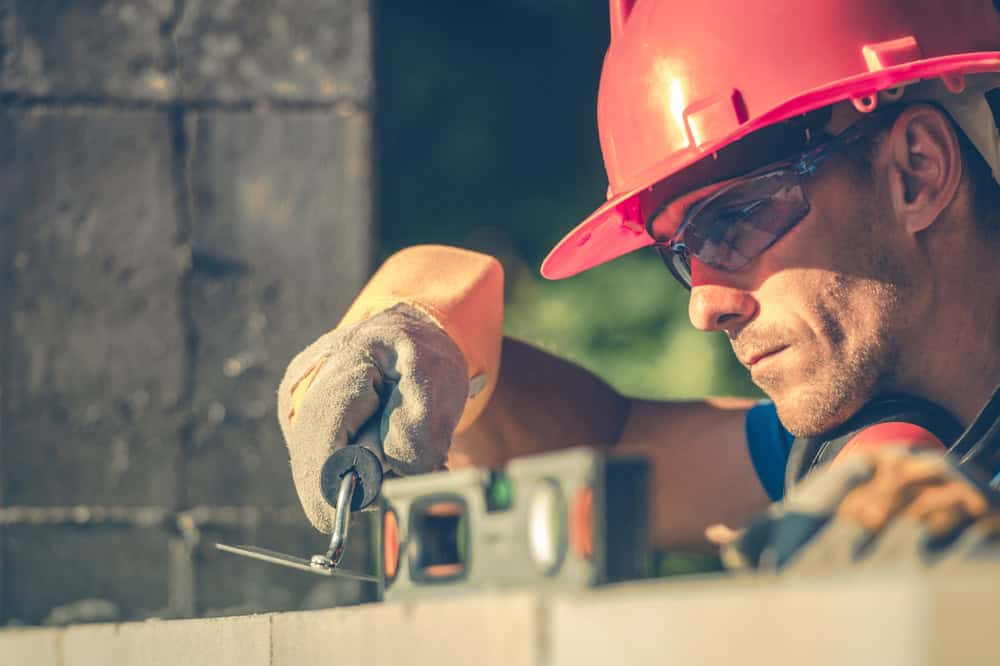 A construction worker wearing a red hard hat, safety glasses, and gloves carefully builds a brick wall, using a spirit level and a trowel to ensure accuracy.