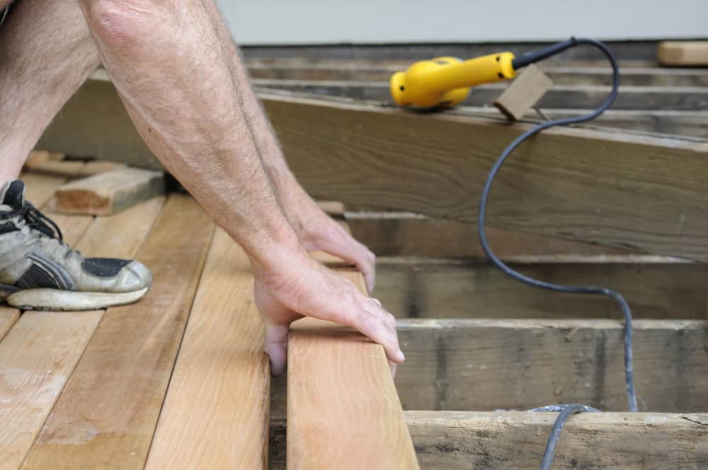 A person wearing sneakers uses both hands to position wooden boards on a deck frame, with a yellow power tool and cord visible nearby.