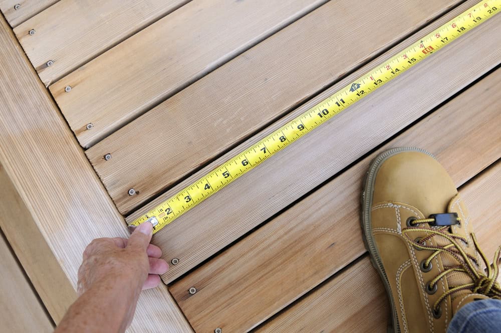 A person measures the width of a wooden deck with a yellow tape measure, while wearing tan work boots and pressing the tape against the boards.