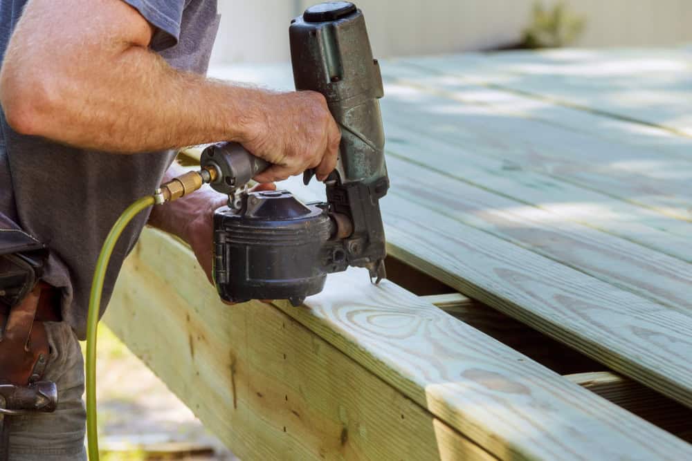 A person uses a nail gun to secure wooden planks while building a deck outdoors. The wood appears new and the worker’s arm is visible, wearing a short-sleeve shirt.