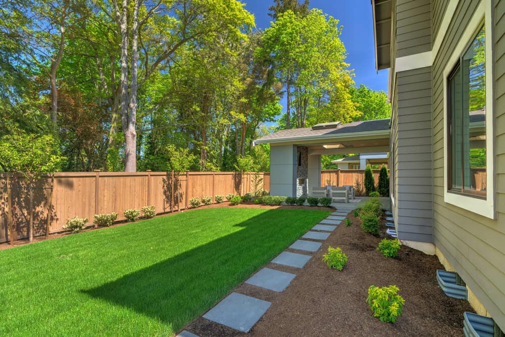 A backyard with bright green grass, a wooden fence, stepping stones leading to a patio area with chairs, and landscaped plants along the edge of a modern gray house on a sunny day.