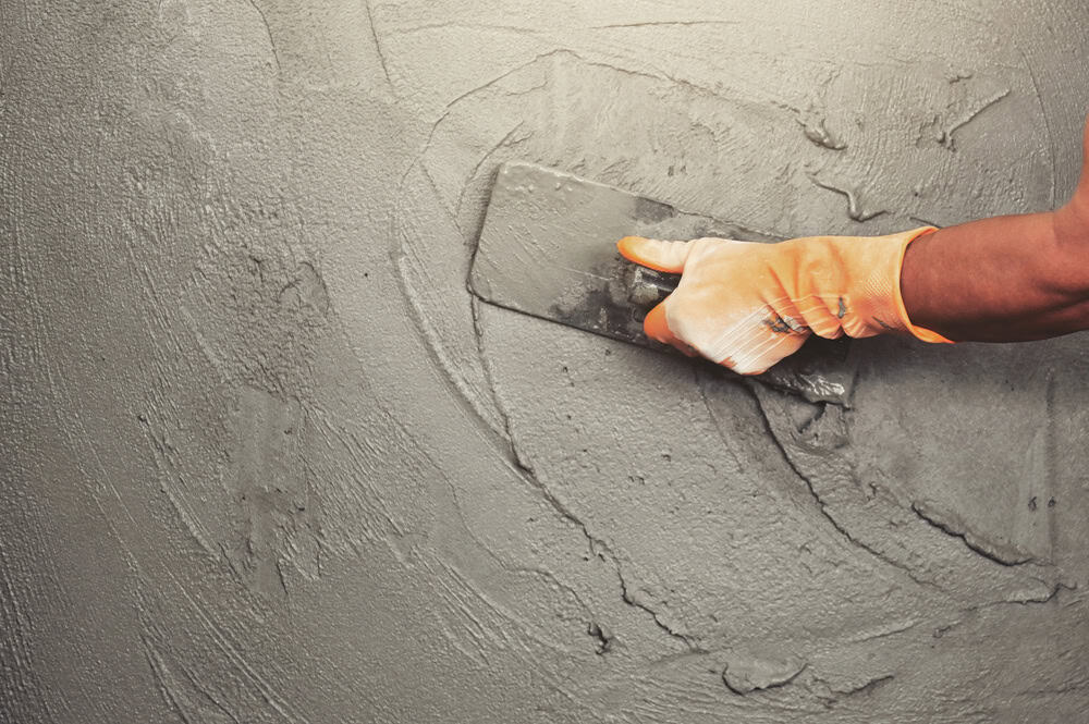 A person wearing an orange glove smooths wet cement on a wall using a trowel.