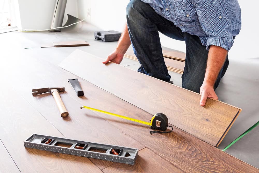 A person installs wood laminate flooring, holding a plank next to measuring tape, spirit level, hammer, and carpenter's square on the floor. The person is wearing a blue shirt and jeans.