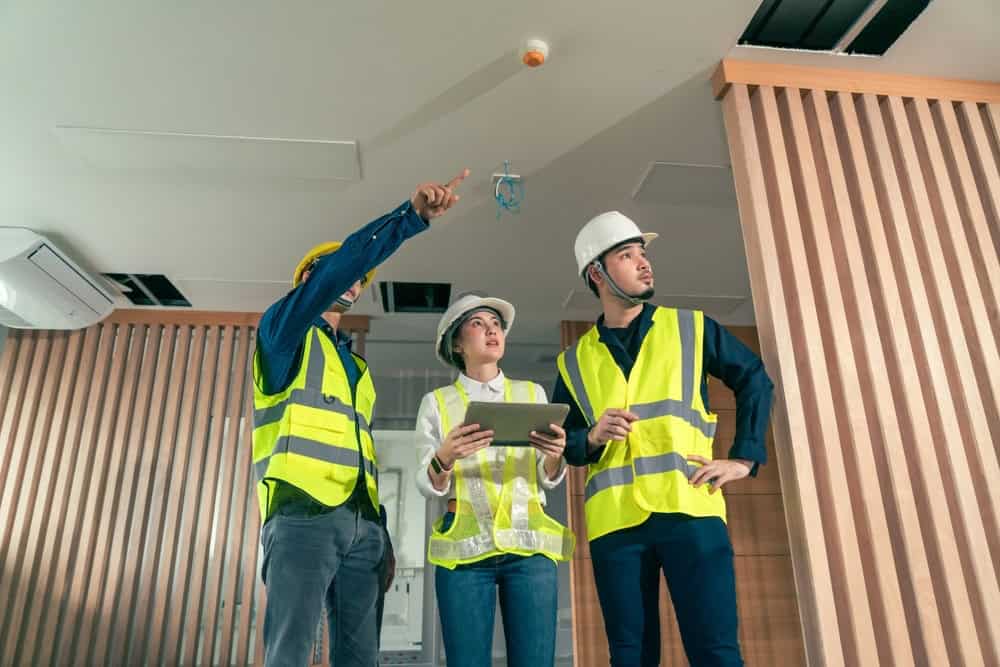 Three construction professionals in safety vests and helmets inspect a modern building interior. One person points upward, another holds a tablet, while the third looks ahead, all observing the ceiling attentively.