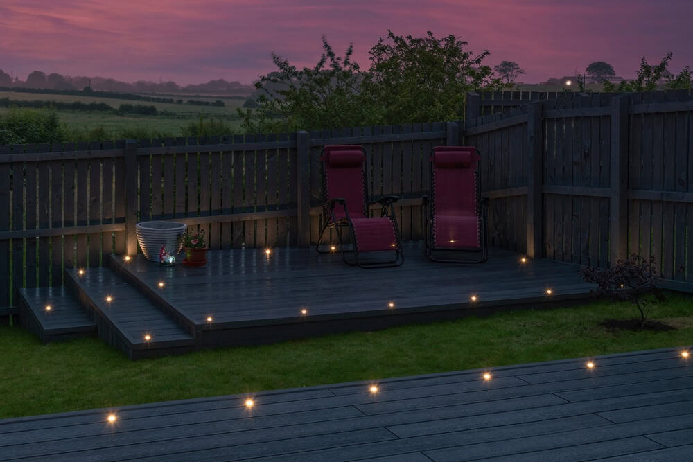 A small, elevated backyard deck with two red reclining chairs, soft deck lights, potted plants, and a gray fence, set against a twilight sky with pink clouds and green fields in the background.