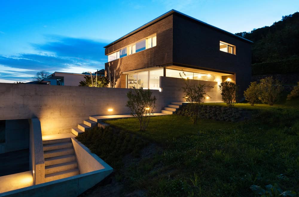 Modern two-story house with large windows and dark exterior walls, illuminated by warm outdoor lights at dusk. Concrete stairs and terraced landscaping lead to the entrance, surrounded by greenery.