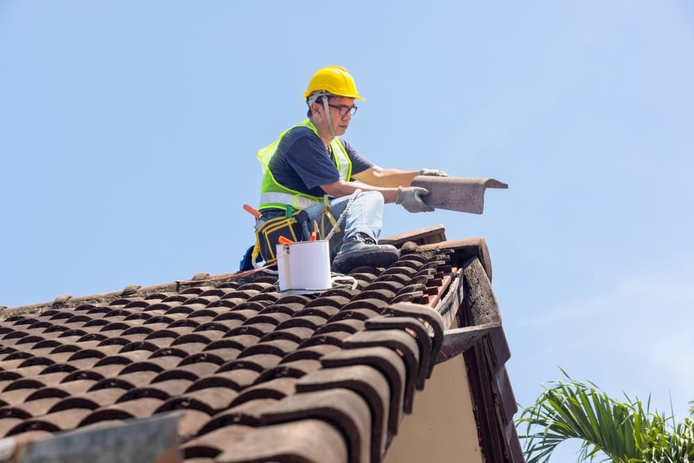 A worker in a yellow hard hat and safety vest repairs a roof, holding a roof tile. A bucket with tools is nearby, and the sky is clear and blue.