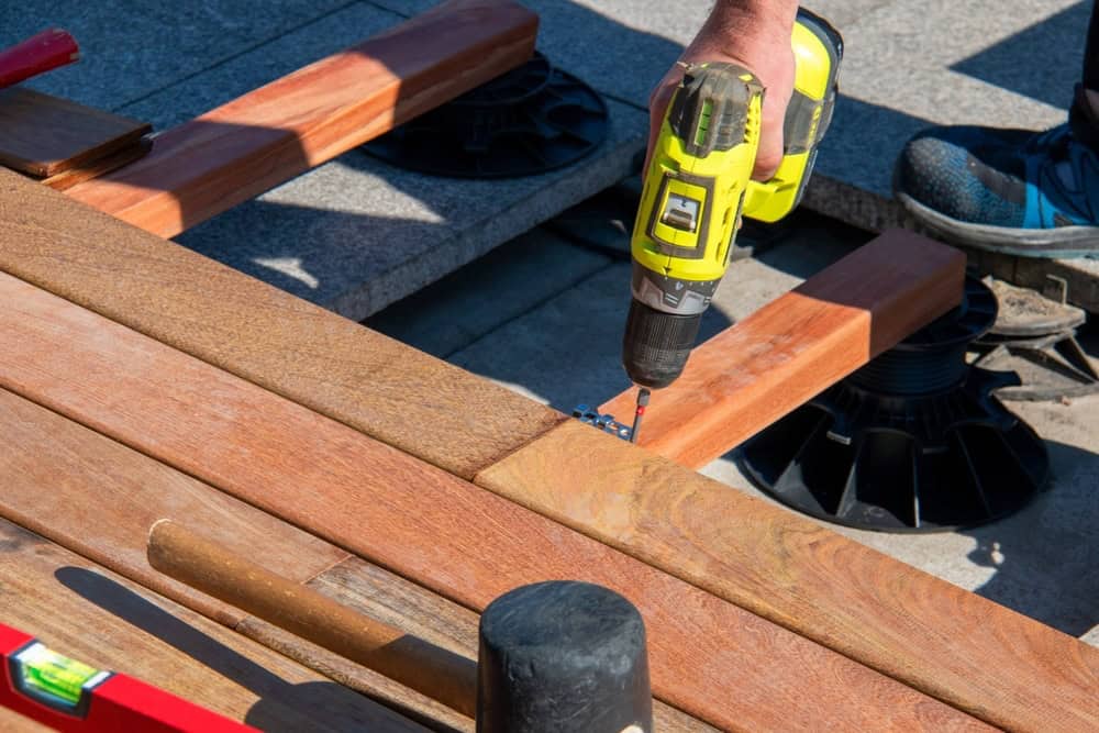 A person uses a yellow cordless drill to fasten wooden planks together on a construction project, with tools and support pads visible nearby.