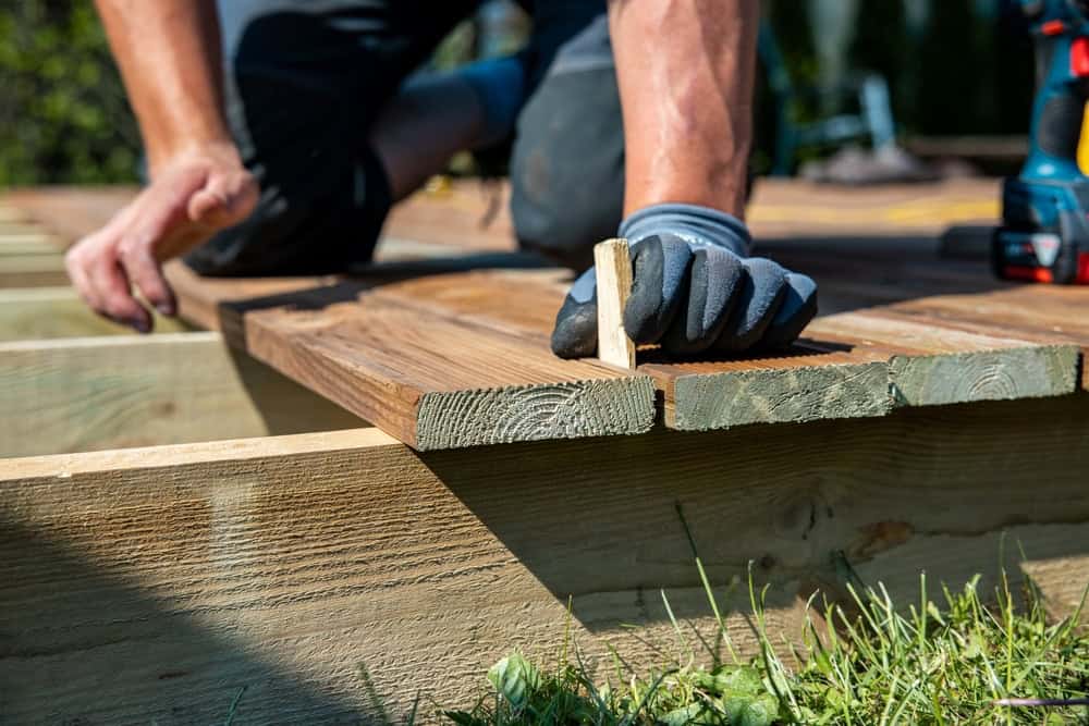 A person wearing gloves installs wooden deck boards outdoors, using a small spacer between boards. Tools and grass are visible in the background.