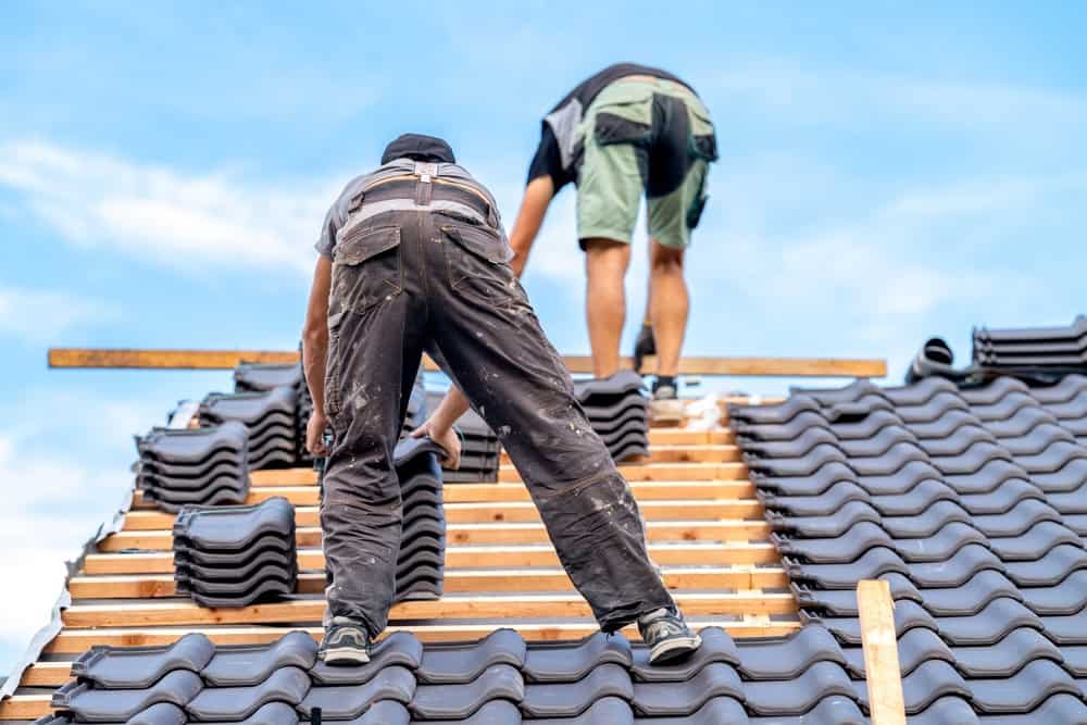 Two workers are installing black roof tiles on a sloped rooftop under a blue sky. They are standing among stacks of tiles and wooden beams, wearing work clothes and safety gear.