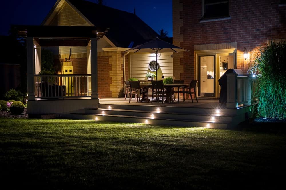 A cozy backyard patio at night features a dining table with chairs under an umbrella, warm deck lighting, a pergola, and well-kept lawn and landscaping beside a brick house.