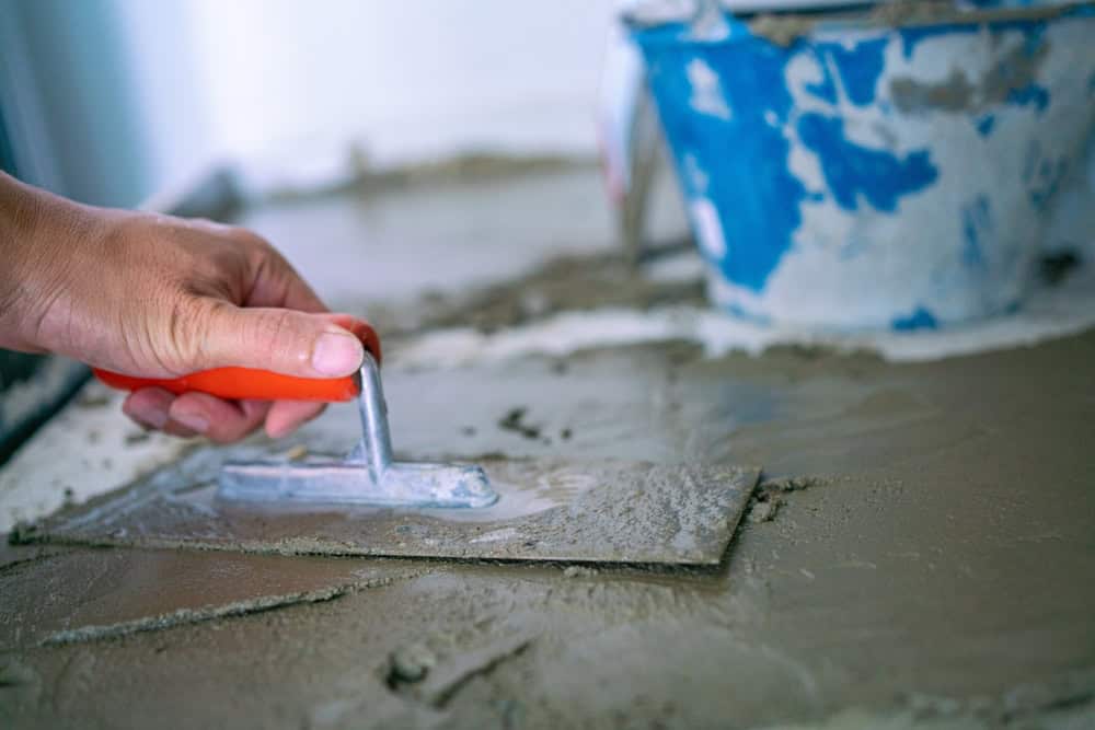 A hand holding a trowel smooths wet cement on a surface, with a blue bucket of cement in the background.