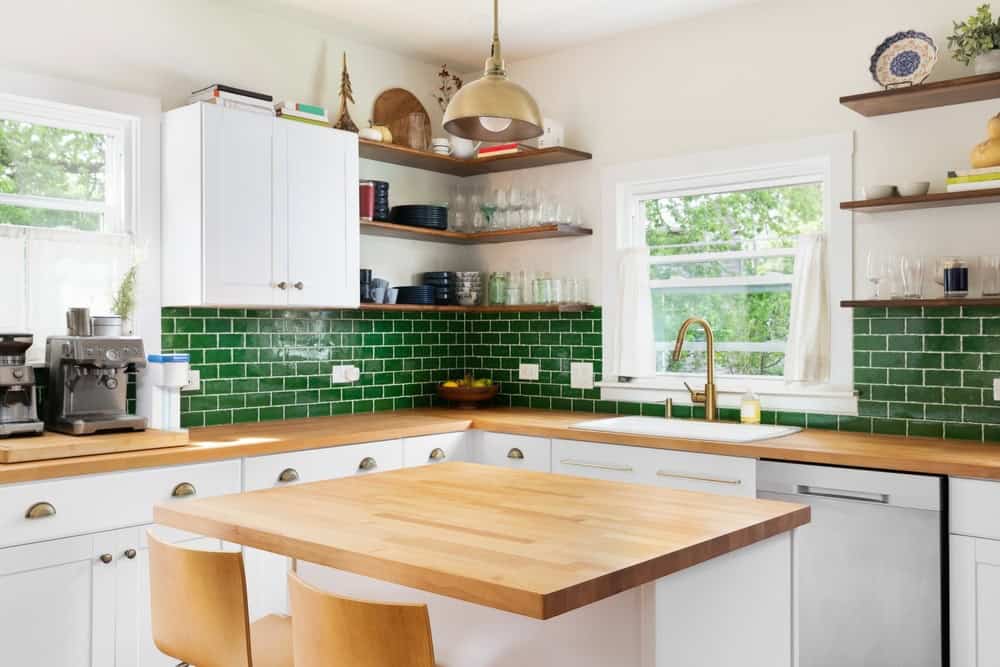 Bright, modern kitchen with white cabinets, wooden countertops, green subway tile backsplash, open shelves with dishes, a coffee machine, and large windows letting in natural light. Two wooden chairs are at a central island.