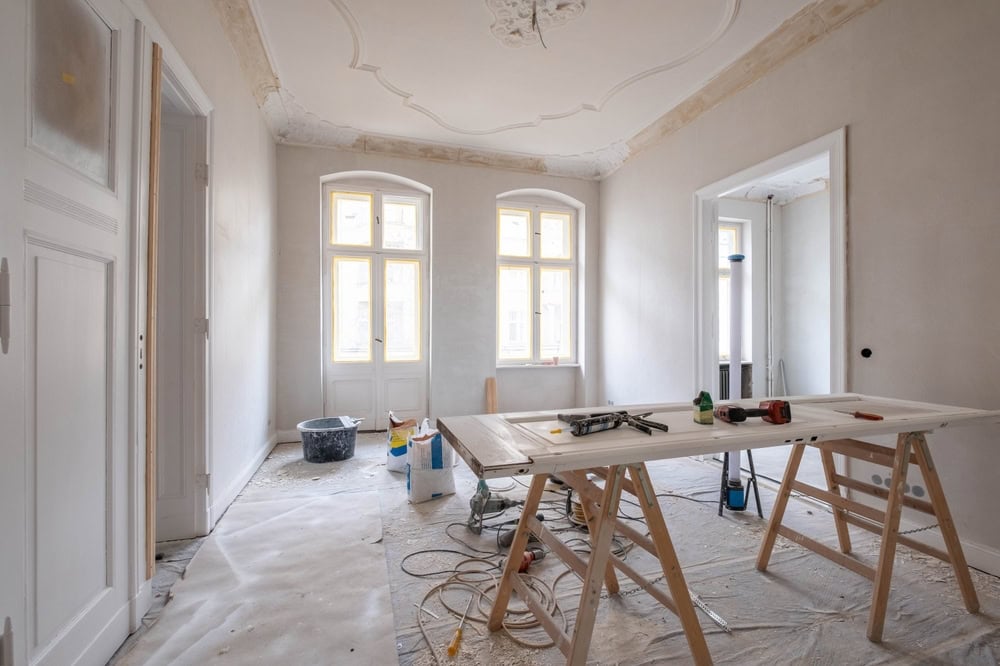 A bright room under renovation with a worktable, tools, and supplies scattered on a plastic-covered floor; windows let in natural light, and the ceiling features decorative molding.