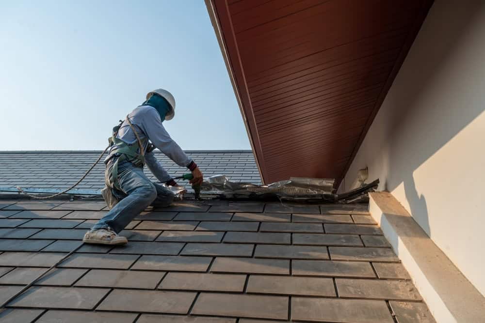 A worker wearing safety gear and a helmet installs or repairs shingles on a sloped roof of a building on a clear day. The worker is using tools near the roof's edge, secured with a safety harness.