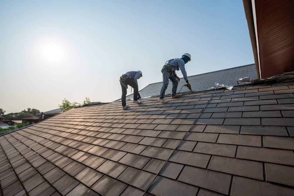 Two workers wearing safety gear install or repair roof tiles on a sloped house roof under bright sunlight, with clear skies in the background.