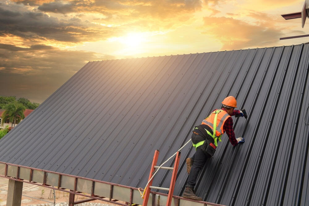 A construction worker wearing a safety harness and helmet installs metal roofing sheets on a building at sunset, with dramatic clouds in the sky.