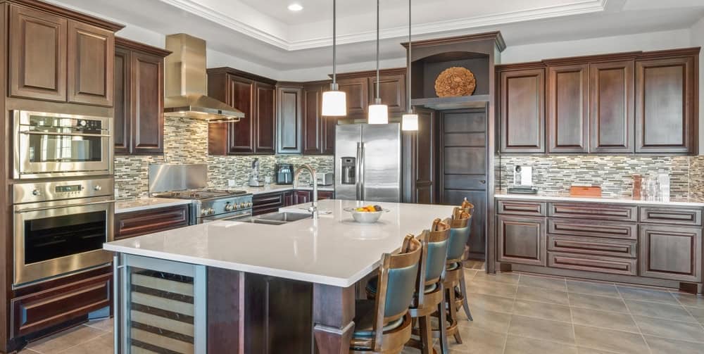 Modern kitchen with dark wood cabinets, stainless steel appliances, a large island with a white countertop, four bar stools, pendant lighting, and a mosaic tile backsplash. A fruit bowl sits on the island.