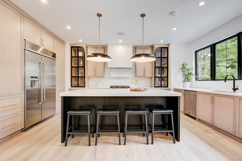 Modern kitchen with light wood cabinets, stainless steel refrigerator, large center island with a white countertop, four black stools, two pendant lights, large window, and built-in oven with range hood.