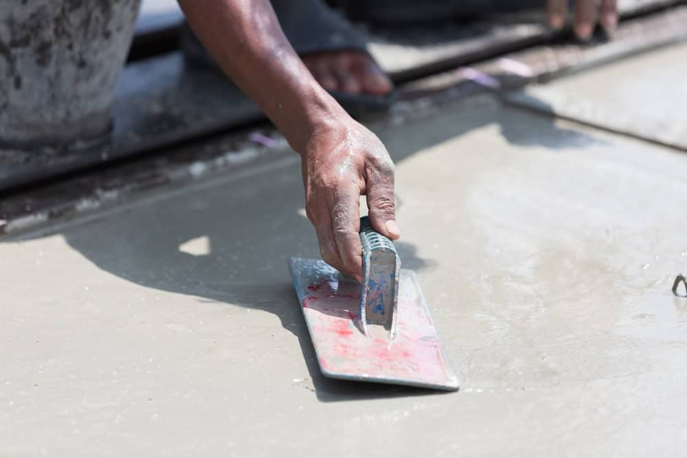 A close-up of a person’s hand using a trowel to smooth wet concrete on the ground. The person’s hand is dirty, and the wet concrete surface is being leveled.