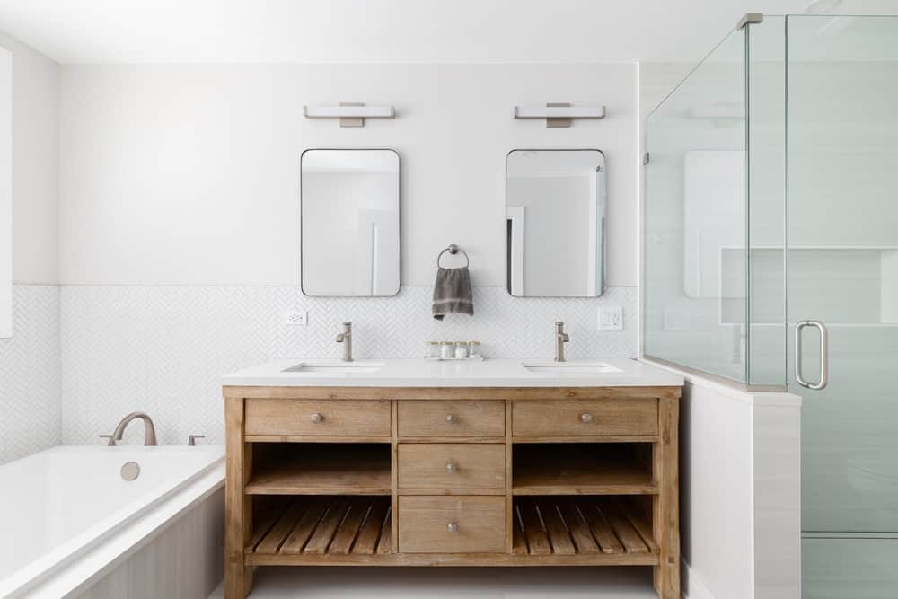 Modern bathroom with a wooden double-sink vanity, two rectangular mirrors, wall-mounted lights, a glass shower enclosure, a bathtub, and white tile walls. A towel hangs between the mirrors.