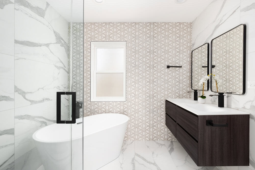 Modern bathroom with a freestanding white bathtub, glass shower door, patterned accent wall, marble-look tiles, double sink vanity with dark wood drawers, two mirrors, and black fixtures.