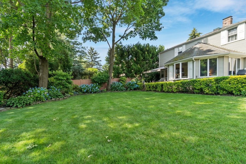 A well-maintained backyard with lush green grass, mature trees, flowering shrubs, and a two-story house with large windows and a sunroom under a clear blue sky.