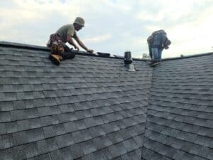 Two workers wearing tool belts are repairing or installing shingles on a sloped roof, with one kneeling and the other bending over near a roof vent under a partly cloudy sky.