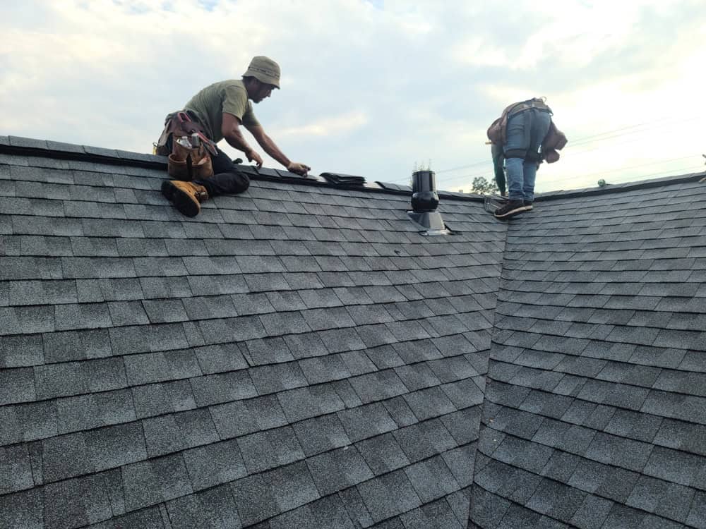 Two workers wearing tool belts are repairing or installing shingles on a sloped roof, with one kneeling and the other bending over near a roof vent under a partly cloudy sky.