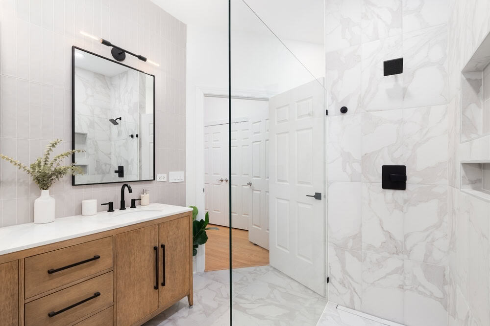 Modern bathroom with a wood vanity, black fixtures, white countertop, a rectangular mirror, light gray tiled walls, a glass shower with marble tiles, and white double doors in the background.