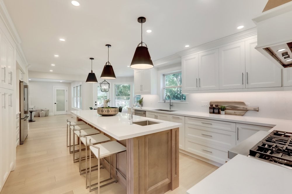 Bright, modern kitchen with white cabinets, a large island with barstools, pendant lights, stainless steel appliances, and light wood floors. Large windows provide ample natural light.
