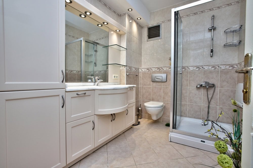 Modern bathroom with white cabinets, large mirror, sink, wall-mounted toilet, glass shower enclosure, and beige tiled walls and floor. Decorative flowers are visible near the door.