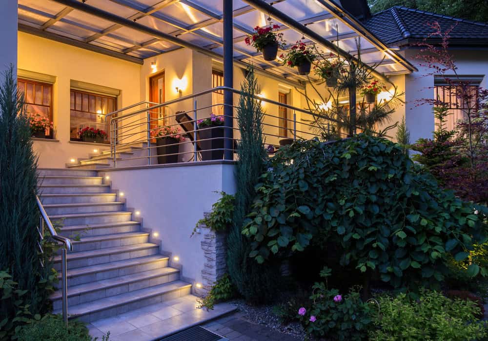 A modern house with a covered, warmly lit porch, metal railings, potted flowers, and steps illuminated by small lights, surrounded by lush green bushes and trees in the evening.