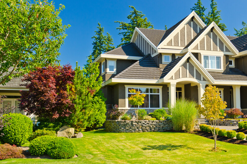 A modern two-story house with a well-maintained lawn, vibrant landscaping, stone retaining wall, and a variety of green trees and shrubs under a clear blue sky.