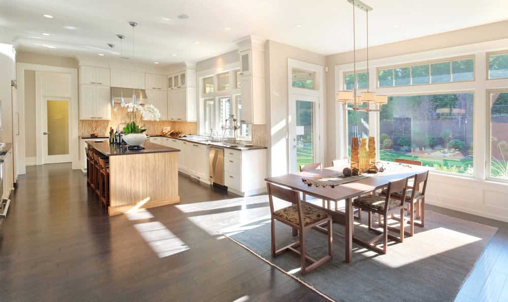 Bright, modern kitchen and dining area with white cabinets, a wood island, large windows, and sunlight streaming in. Dining table with chairs sits on a rug, overlooking a green backyard through tall windows.