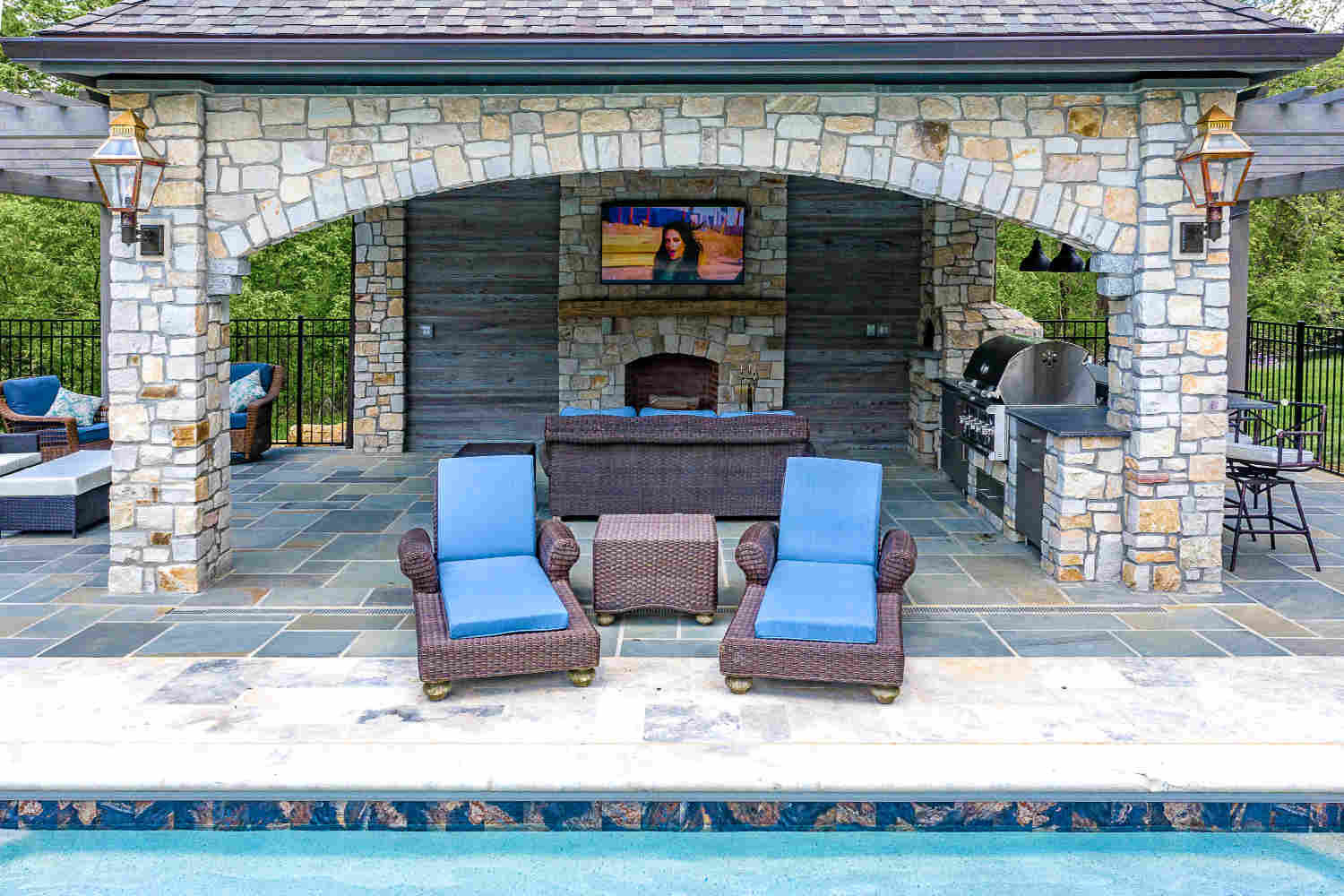 Outdoor poolside lounge area with two blue-cushioned wicker chairs and a table facing a stone pavilion, which features a TV, built-in grill, bar seating, and wooden accents.