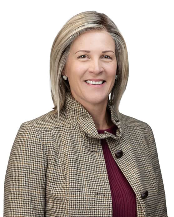A woman with short blonde hair smiles, wearing a brown houndstooth jacket over a maroon top. She poses in front of a plain white background.