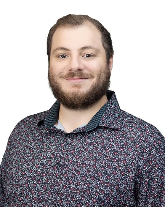 A bearded man with short hair smiles at the camera, wearing a dark patterned button-up shirt with a white background.