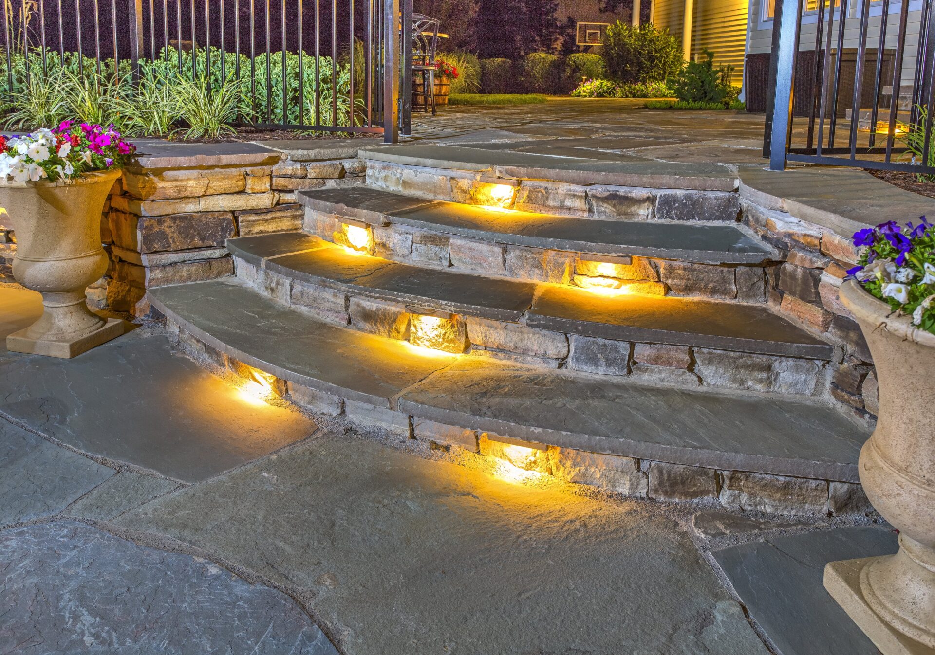 Curved stone steps with built-in yellow lights lead to a patio area. Potted flowers sit on either side of the steps, and a garden with plants and grass is visible in the background at night.