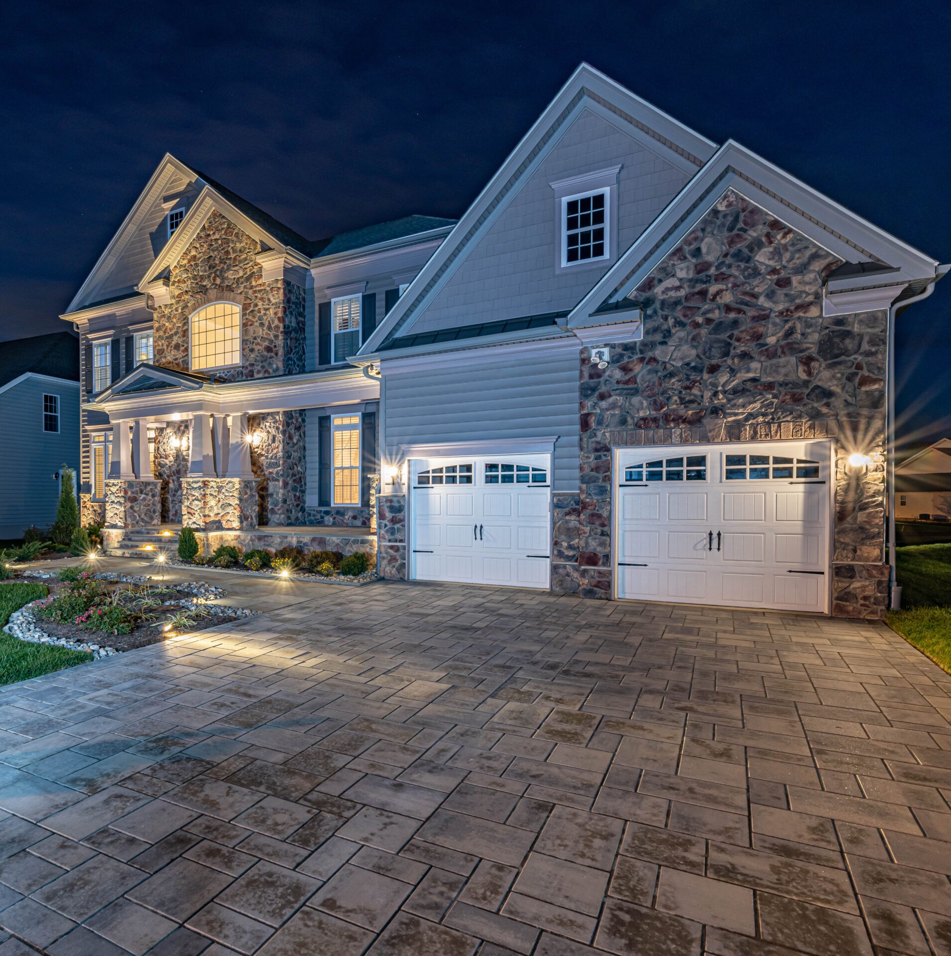A large two-story house with stone and siding exterior, illuminated by outdoor lighting at night. The home features two garage doors, a paved driveway, and landscaped garden beds.